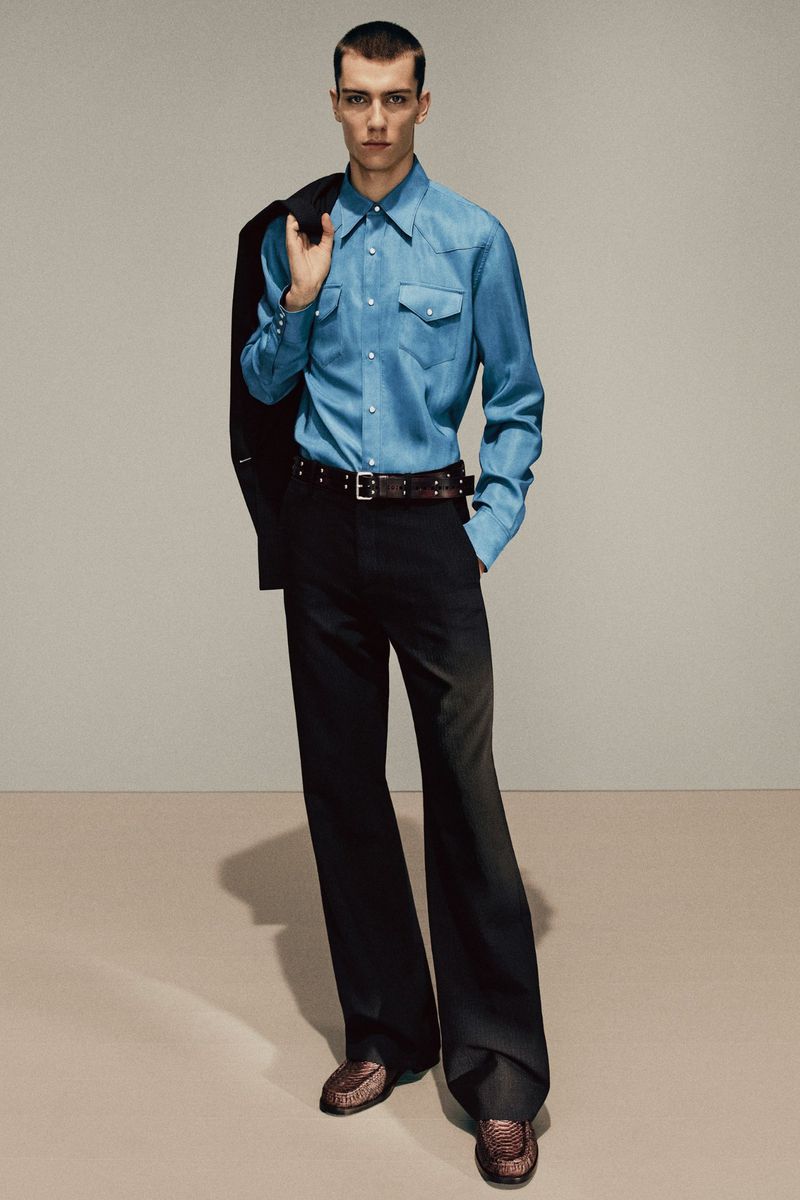 A full-length portrait of a slender adult male wearing a Western-style denim shirt, dark trousers, and dress shoes, posing in a studio.