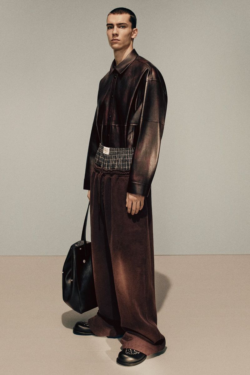 Model wearing a dark brown leather shirt and wide trousers holding a black handbag against a grey studio backdrop.