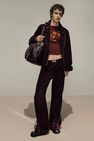 Model in dark striped velvet jacket and trousers holding a black leather bag against a neutral studio background