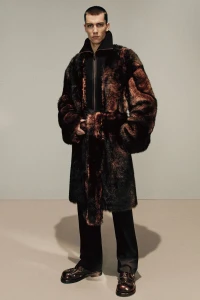 Model wearing a dark faux-fur coat and black trousers against a neutral grey studio background for Acne Studios
