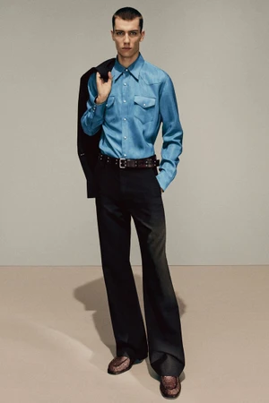 Male model in blue button-down shirt and black trousers holding a jacket against a neutral studio background.