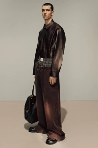 Model wearing a dark brown leather shirt and wide trousers holding a black handbag against a grey studio backdrop.