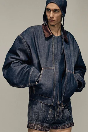 Male model wearing a dark denim jacket with contrast stitching, matching shorts, and a matching cap against a neutral grey studio background