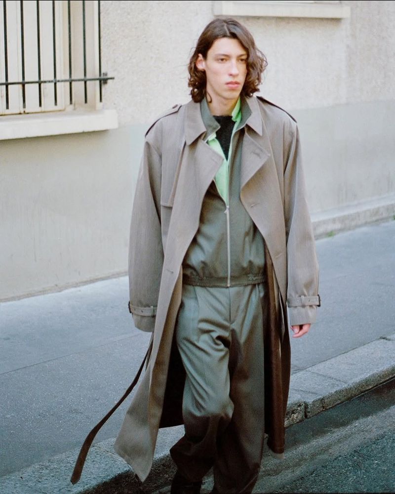 An adult male strides down a sidewalk in a light beige trench coat, green tracksuit, and black shoes, next to a light building with dark bars.