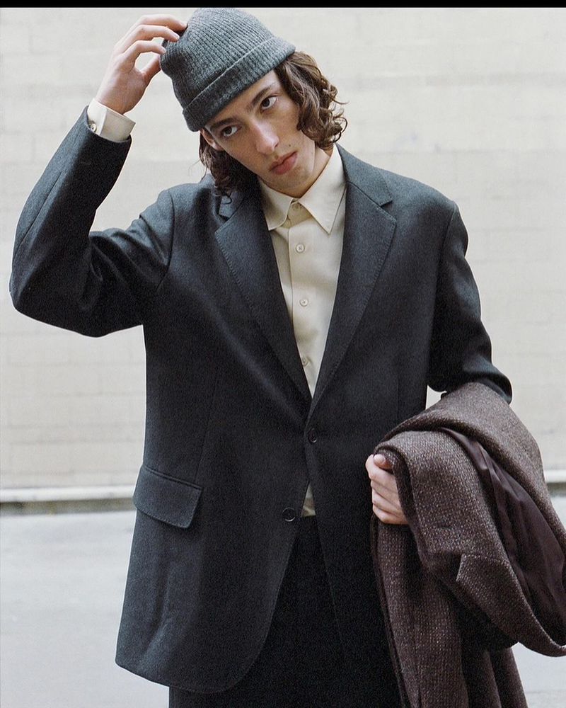 Young man wearing a charcoal blazer, cream shirt, and grey beanie while holding a brown coat outdoors
