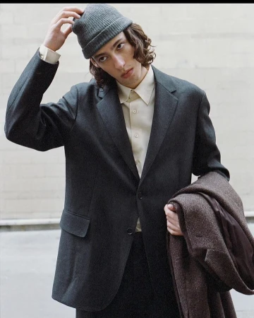 Young man wearing a charcoal blazer, cream shirt, and grey beanie while holding a brown coat outdoors
