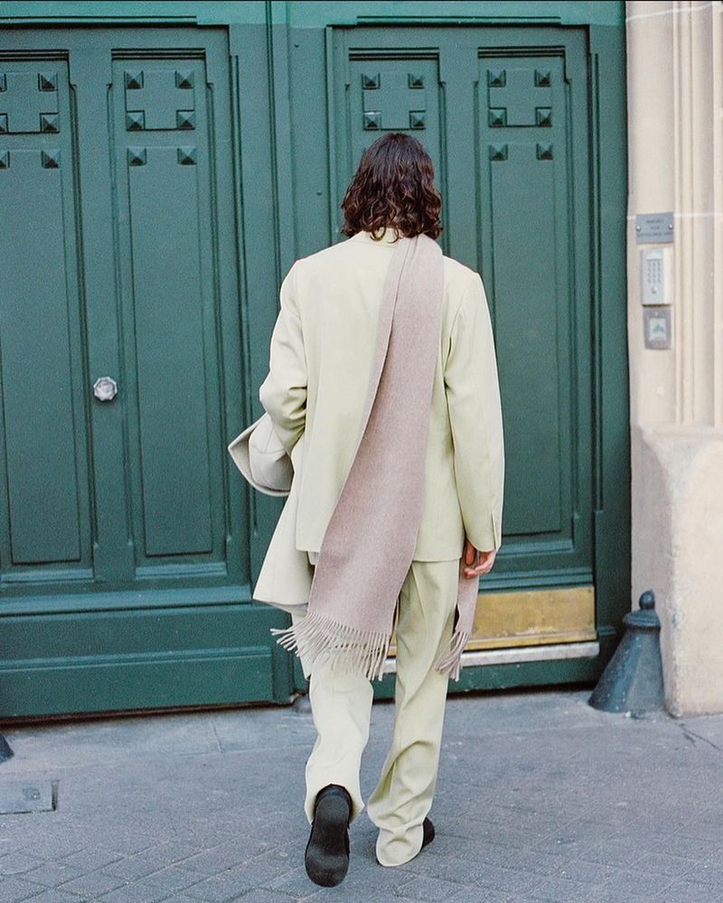 Back view of a slender person wearing a cream suit and brown scarf walking away on a sidewalk in front of a green door.