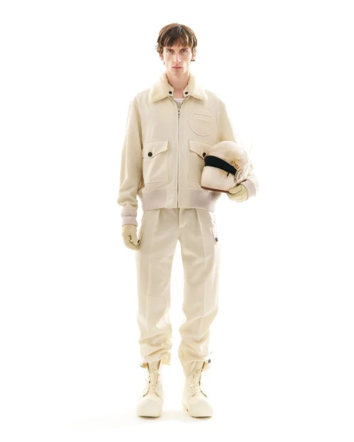 Full-shot of a person in an off-white jacket, pants, and boots. They are holding a racing helmet in a brightly lit studio.