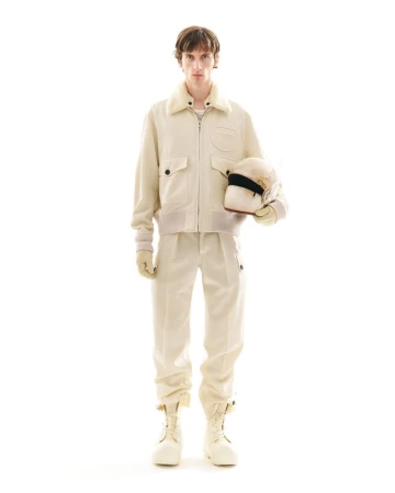 Full-shot of a person in an off-white jacket, pants, and boots. They are holding a racing helmet in a brightly lit studio.