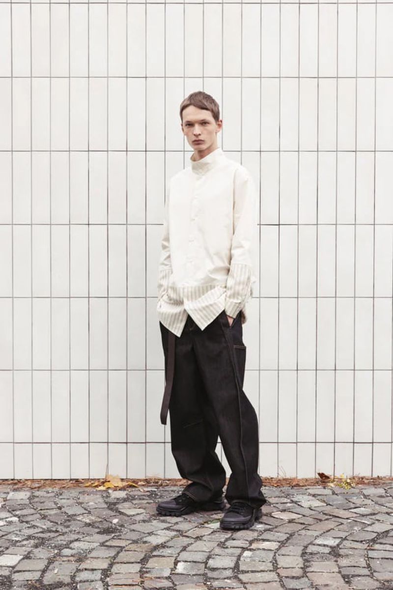 Full shot of a male model in an off-white shirt and black pants posing against a white tile wall with a cobblestone ground outdoors.