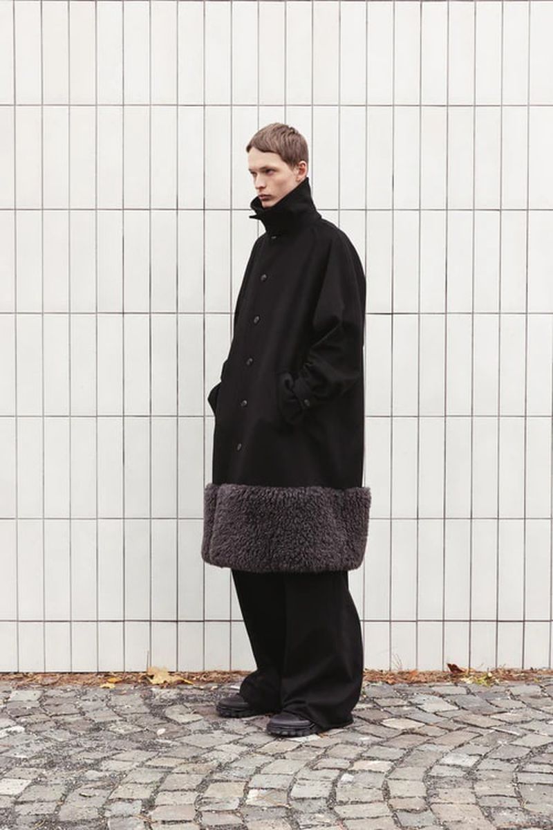 A slender adult male poses in a long, dark wool coat with a light-grey faux fur hem. He wears wide-legged dark pants and chunky black shoes.