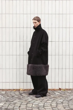 A slender adult male poses in a long, dark wool coat with a light-grey faux fur hem. He wears wide-legged dark pants and chunky black shoes.