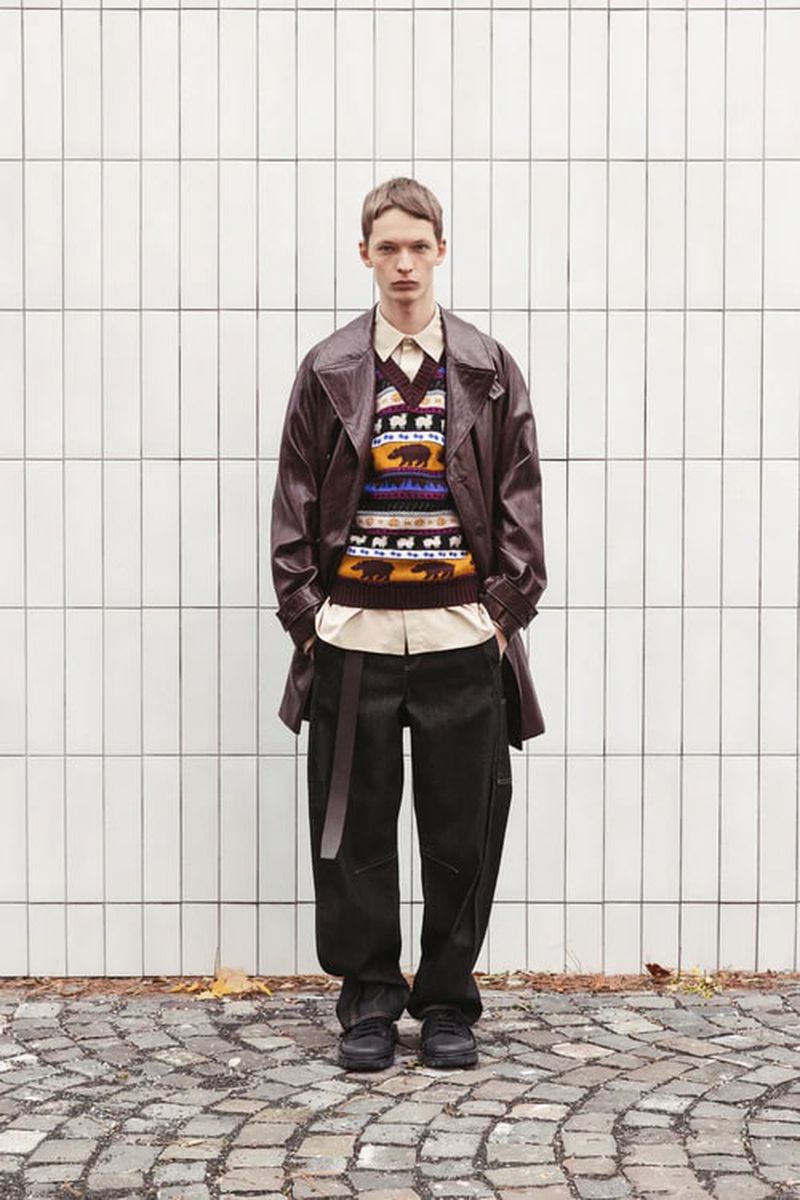 Male model in a brown trench coat, patterned sweater vest, and dark pants stands against a white tiled wall on a cobblestone path.