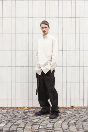 Model David Kenvesh wearing an off-white shirt and dark trousers standing against a tiled wall