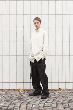 Model David Kenvesh wearing an off-white shirt and dark trousers standing against a tiled wall