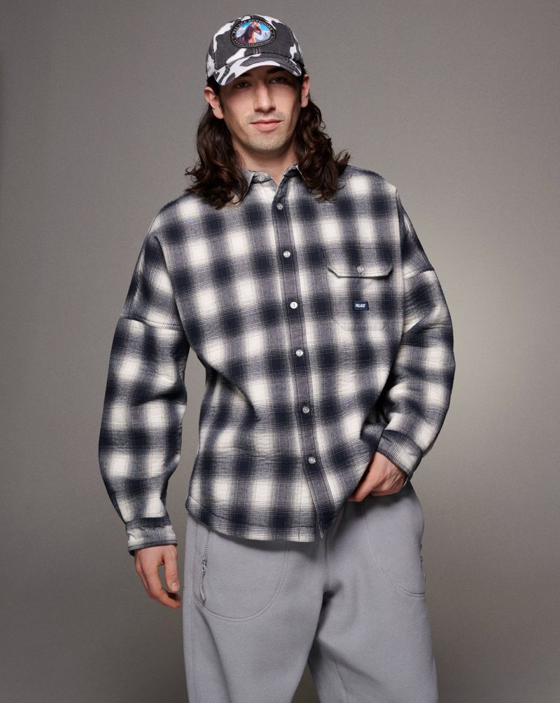 Male model poses indoors wearing an oversized plaid shirt, gray pants, and a patterned cap, set against a neutral background in a studio shot.