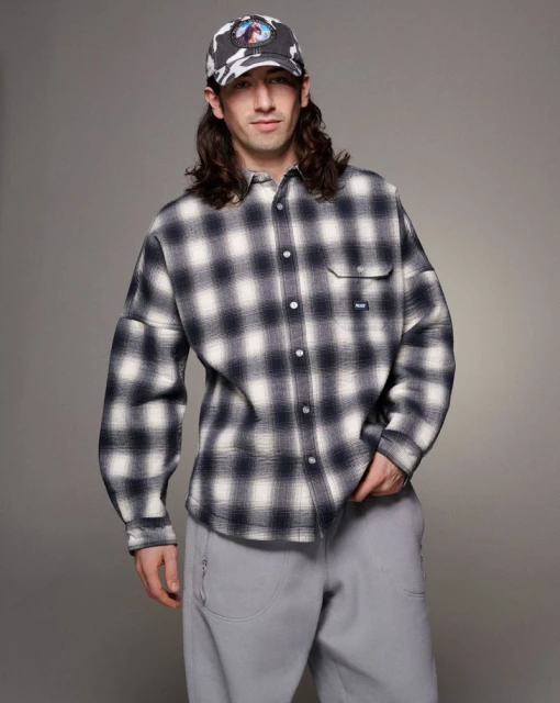 Male model poses indoors wearing an oversized plaid shirt, gray pants, and a patterned cap, set against a neutral background in a studio shot.