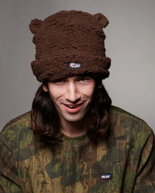 Young adult wearing a brown furry hat with bear ears and a camouflage shirt, smiling in a studio portrait with soft lighting.