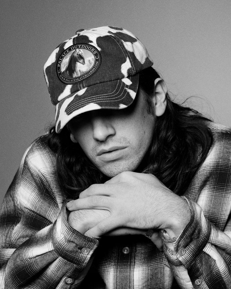Black and white portrait of a person with long dark hair wearing a cap and flannel shirt looking down in an indoor setting.