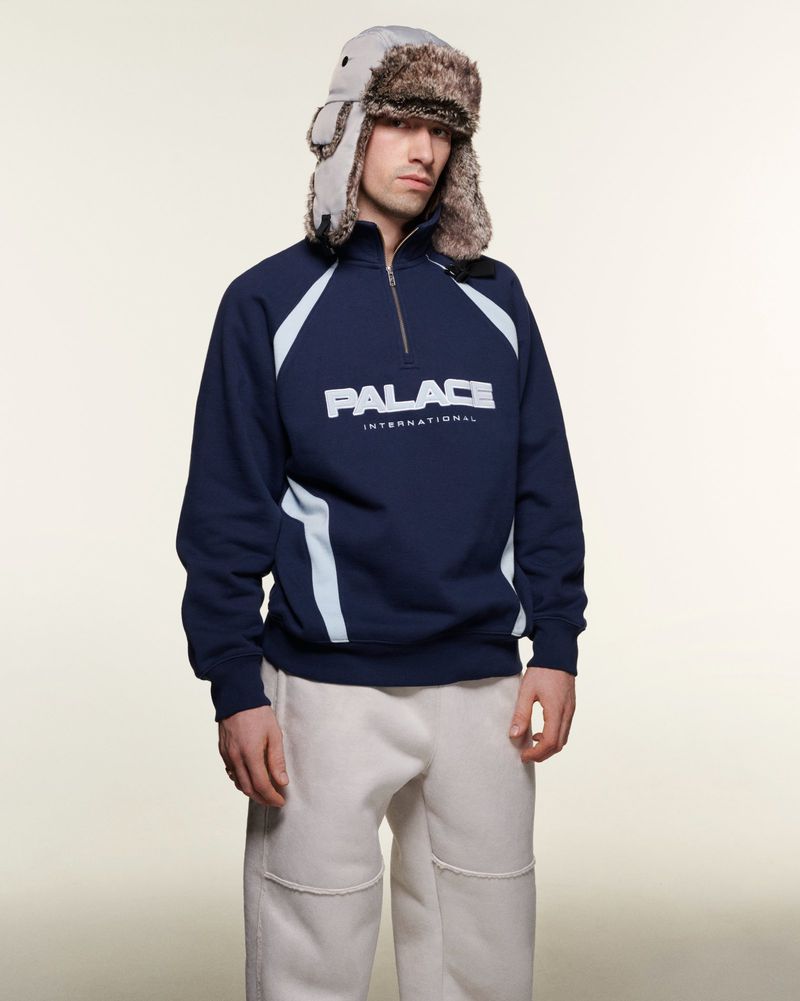 A fashionable adult male stands indoors wearing a navy sweatshirt with white accents, paired with light-colored pants and a fur-trimmed hat.