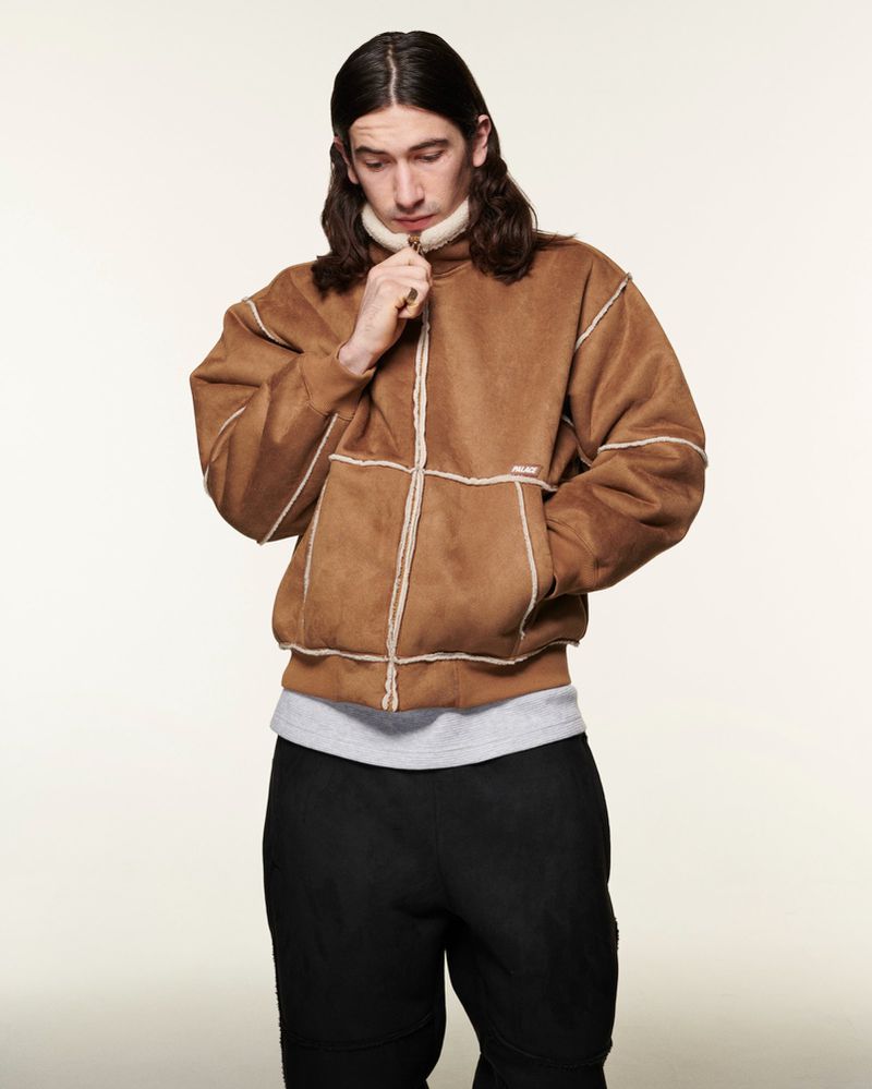Adult male in a brown suede jacket with white trim and black pants stands indoors against an off-white backdrop, looking down.