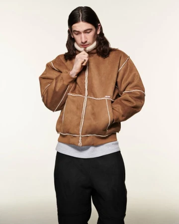 Adult male in a brown suede jacket with white trim and black pants stands indoors against an off-white backdrop, looking down.