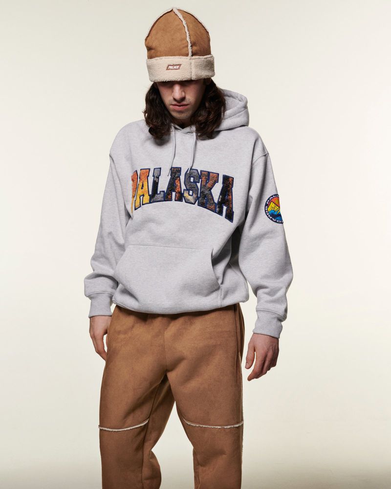 Young adult wearing a gray Alaska hoodie, brown pants, and a shearling hat posing indoors in a studio with a neutral backdrop.