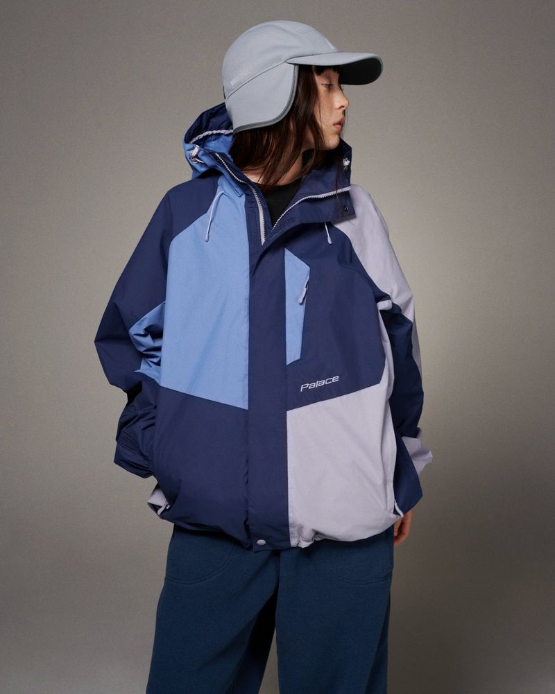 Woman in a blue and grey color-block hooded winter jacket and grey cap with ear flaps poses in a studio. Fashion shot, neutral background.