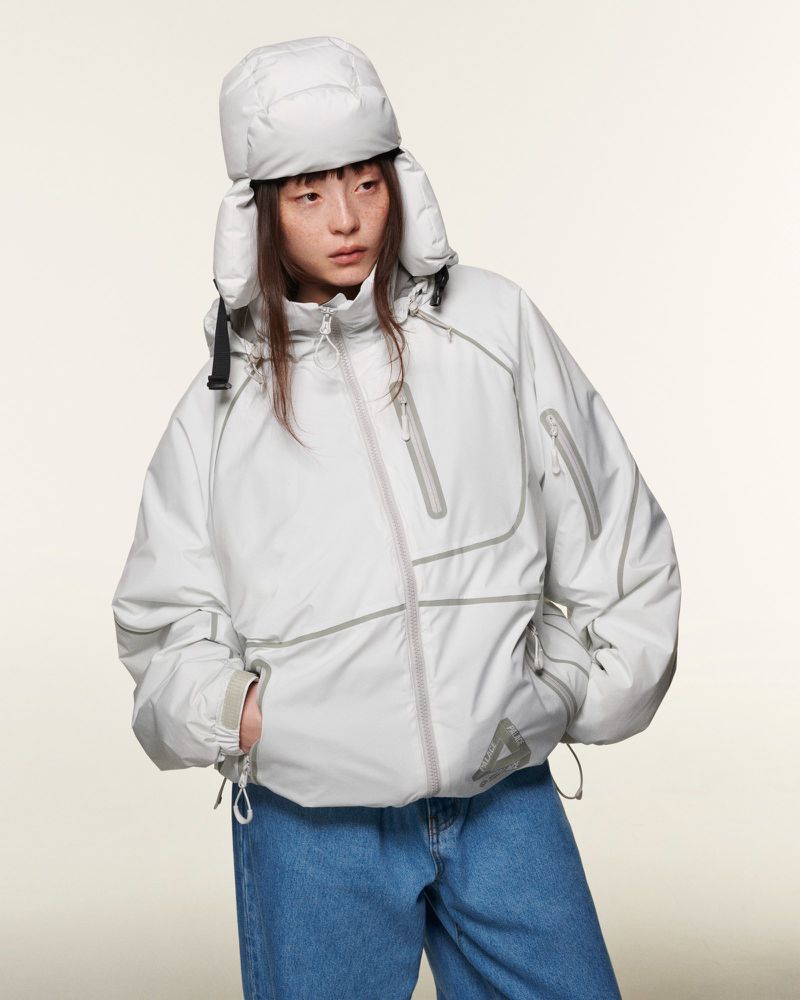 A woman in a white puffer jacket and trapper hat poses indoors. The jacket has reflective accents and the background is neutral.