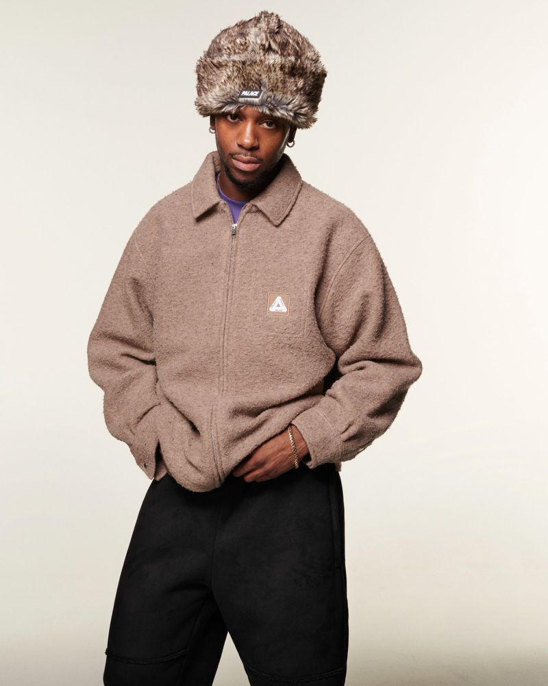 A young man in a fur hat and brown jacket poses indoors against a white backdrop, hands near his waist, in a fashion shot.
