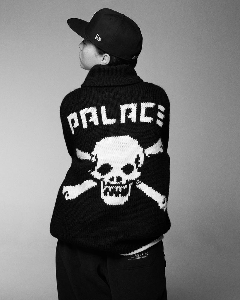 Person wears a black baseball cap and black knitted sweater with a skull and crossbones graphic facing away from the camera in a studio shot.