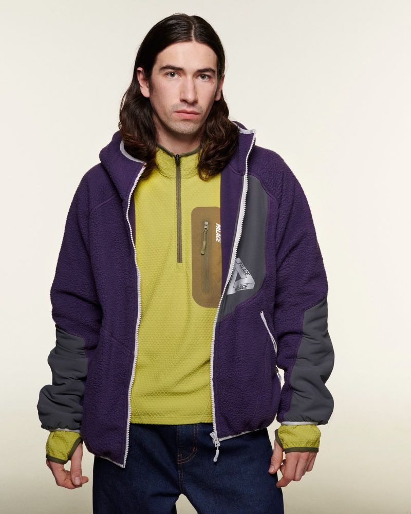 Man in a purple fleece zip-up jacket and yellow shirt posing against a white background in a studio for a fashion shot.