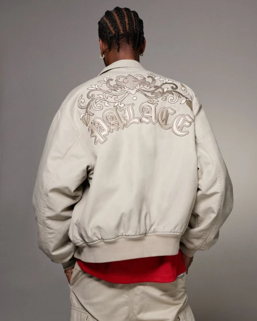 Man wearing a tan Palace jacket with embroidered back design and tan pants against a neutral background in a studio setting.