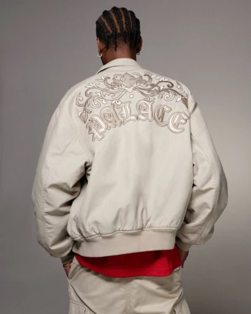 Man wearing a tan Palace jacket with embroidered back design and tan pants against a neutral background in a studio setting.