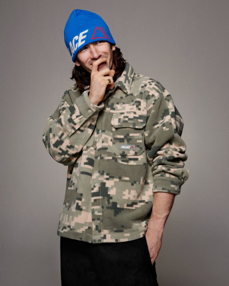 Man in camo shirt and blue beanie poses indoors with hand near face, showcasing casual style against neutral background.