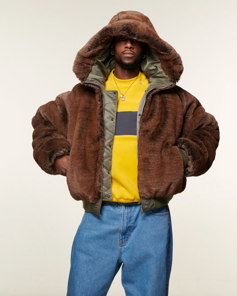 A Black man wearing a brown faux fur jacket with a hood, a yellow striped shirt, and blue jeans poses against a neutral backdrop.
