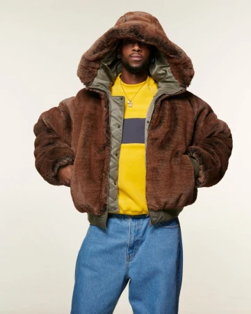 A Black man wearing a brown faux fur jacket with a hood, a yellow striped shirt, and blue jeans poses against a neutral backdrop.