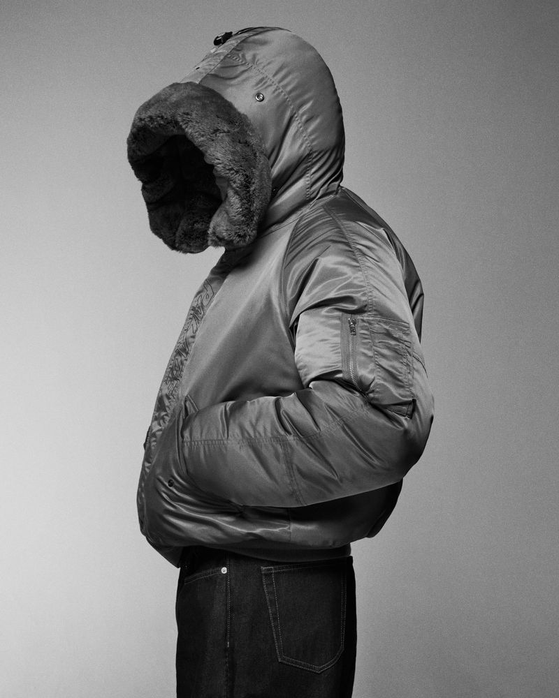 Person wearing a puffer jacket with a fur-trimmed hood in a studio shot. The individual is dressed in dark jeans against a grey background.