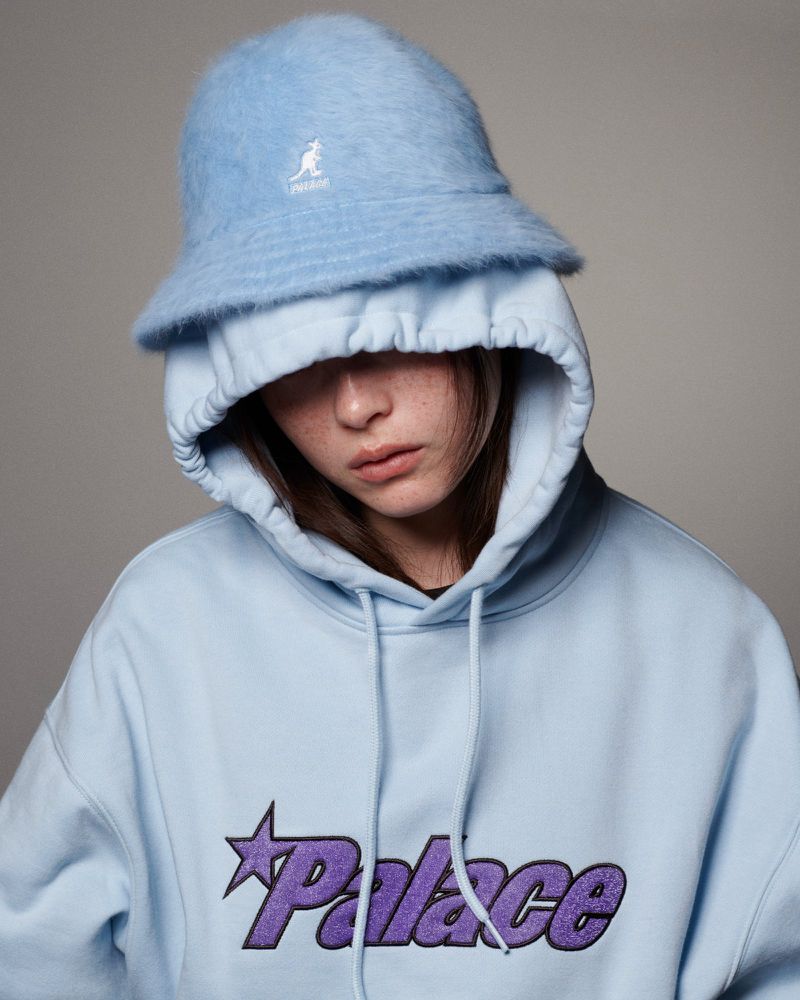 A young woman in a light blue Palace hoodie and fuzzy Kangol bucket hat poses in a studio. Fashion and youth culture are visible.