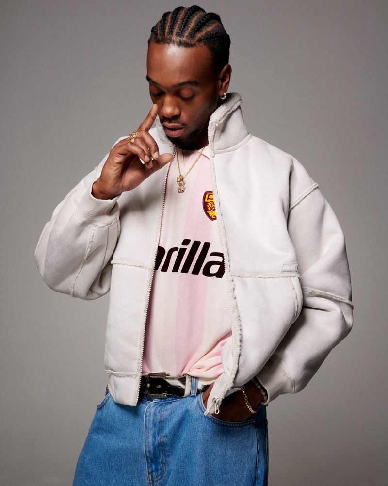 Stylish man in a white jacket, blue jeans and black belt, looking down and posing in a studio with a grey background. 