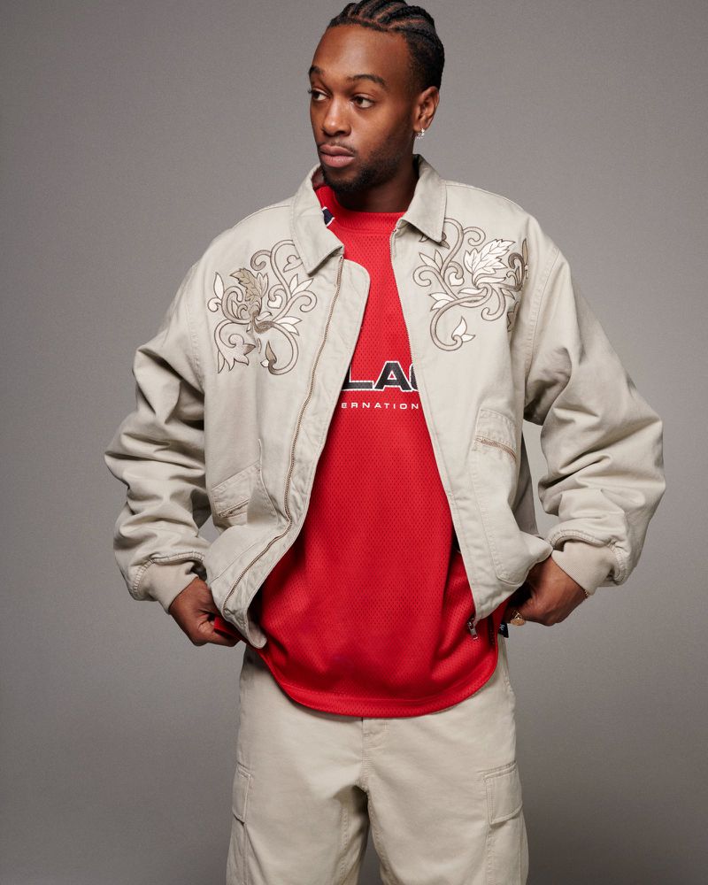 Man in tan jacket and pants stands in a photo studio, looking to the side. He wears a red jersey under his jacket.