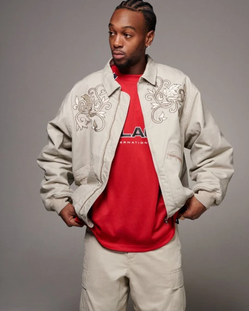 Man in tan jacket and pants stands in a photo studio, looking to the side. He wears a red jersey under his jacket.