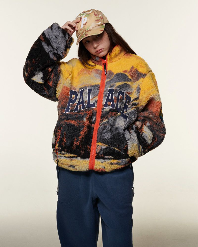 Woman wearing a patterned fleece jacket and cap stands indoors. The fleece has yellow and grey patterns with the 'Palace' logo. 