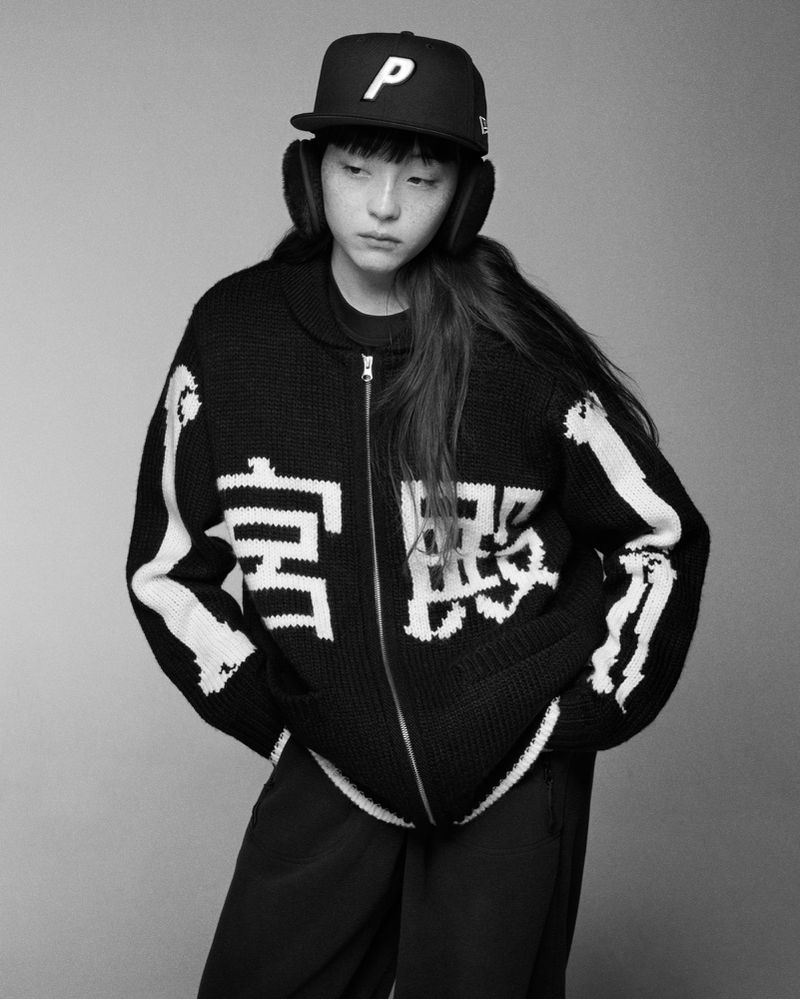 Portrait of a young adult female wearing a graphic zip-up sweater, baseball cap, and earmuffs in a monochrome photo studio shot.