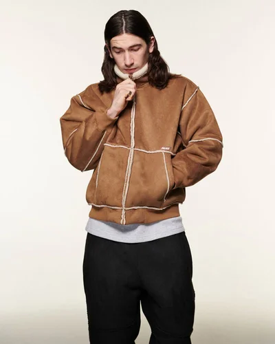 Man with long dark hair wearing a brown shearling jacket zipping up his collar against a seamless light background