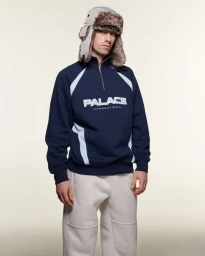 Model wearing a navy blue quarter-zip sweatshirt, white sweatpants, and a grey furry trapper hat against a plain background.