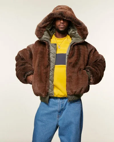 Man wearing a brown faux fur hooded jacket over a yellow sweater and blue jeans against a neutral background.