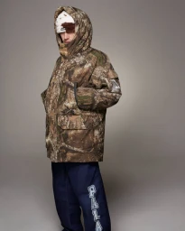 Model wearing a camouflage hooded jacket and blue pants against a neutral grey studio background for PALACE Holiday 2024.
