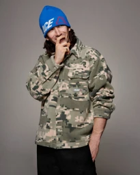 Man wearing a blue beanie and camouflage fleece jacket standing against a neutral grey studio background