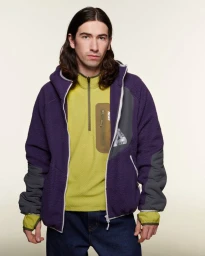 Long-haired man wearing a purple fleece zip jacket over a yellow half-zip fleece and dark jeans against an off-white background